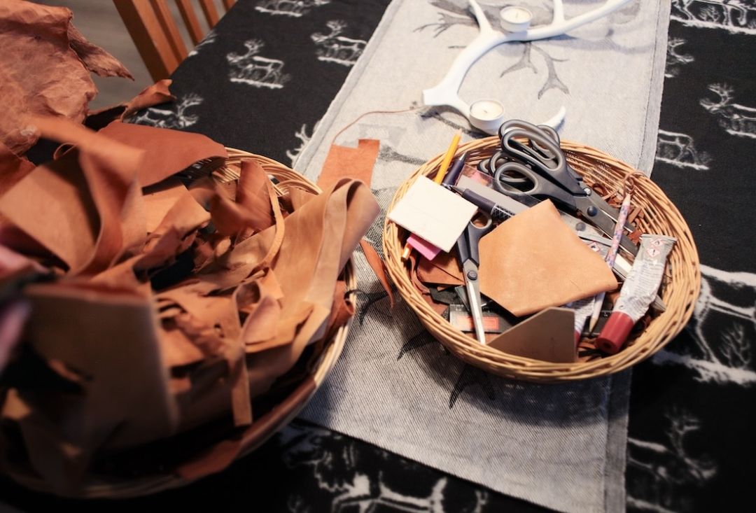 Askartelutarvikkeita, kuten sakset ja nahkanpaloja korissa pöydällä olevan pöytäliinan päällä. Kuva Miika Leivo