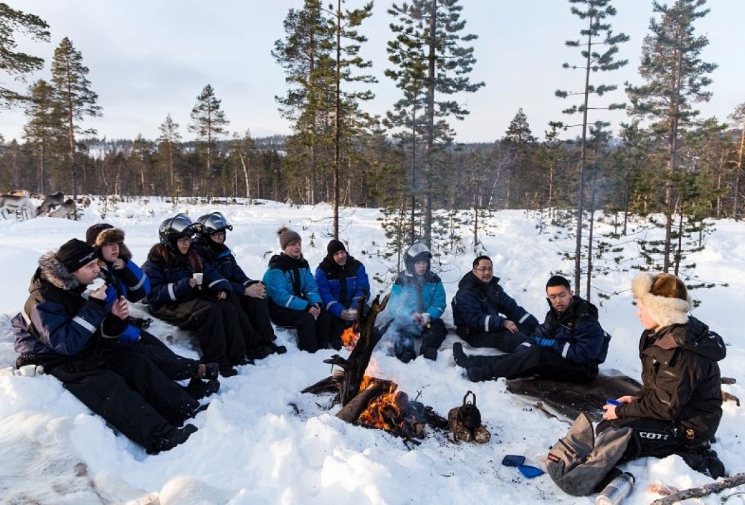 Retkeilijäjoukkio istuu lumessa nuotion ympärillä.