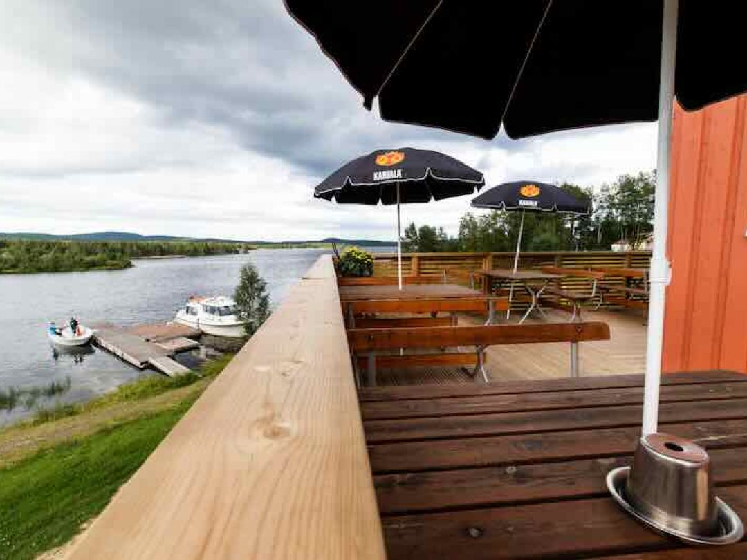 Ulkoterassi puisine penkkeineen ja aurinkovarjoineen, järvi ja veneitä taustalla.