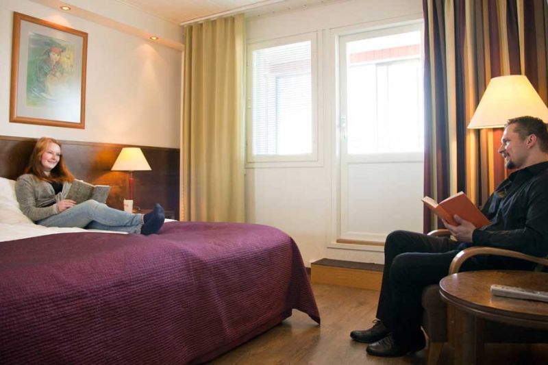 Nainen makoilee parisängyllä, joka on ikkunan lähellä. Mies lukee kirjaa nojatuolissa sängyn päädyssä.