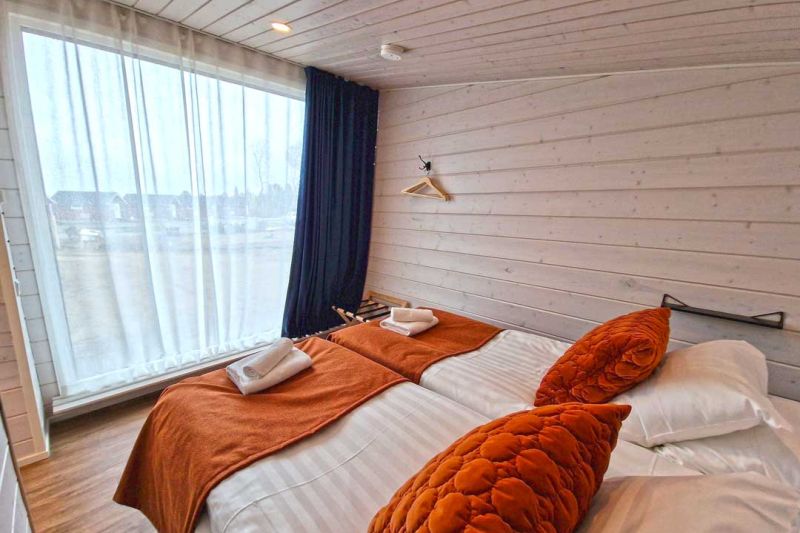 Puupaneelinen Sauna Studion hotellihuone suurella ikkunalla. Rauhallinen tunnelma vaalealla värimaailmalla, jota on piristetty lämpimällä oranssin sävyllä.