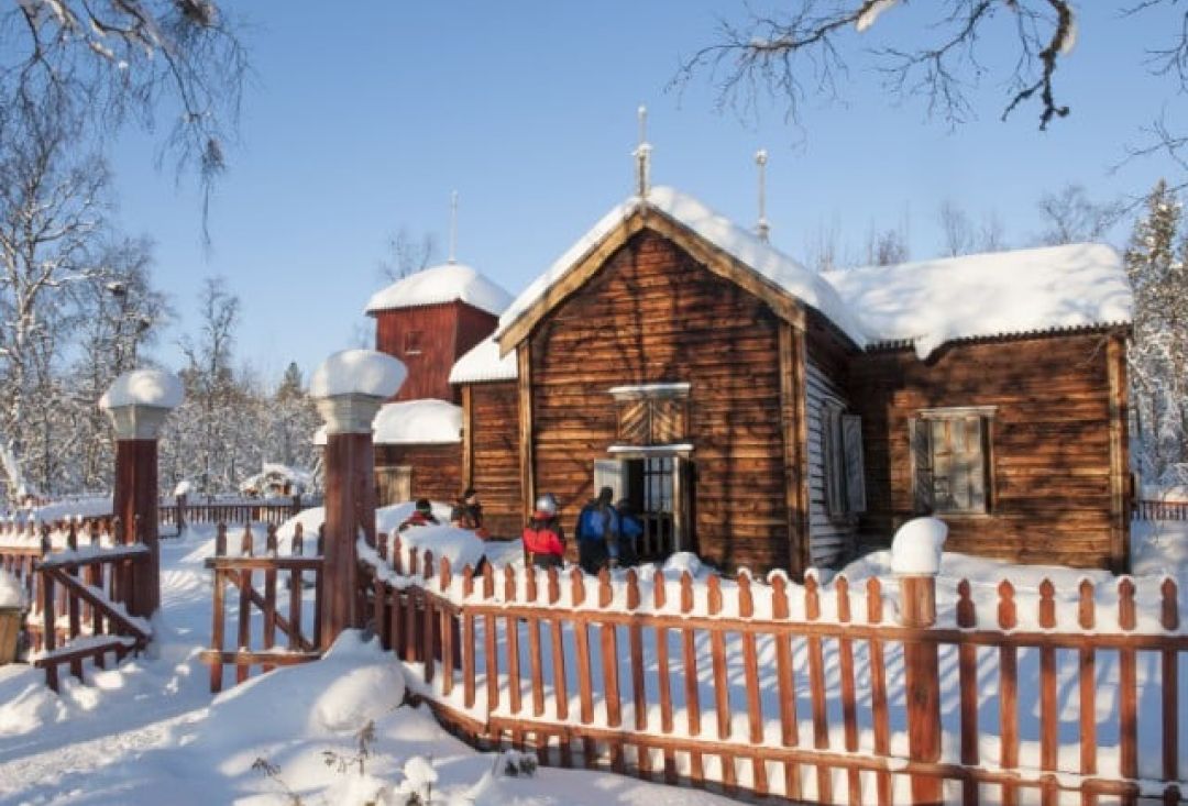 Pielpajärvi church in snowy scenery, surrounded by wooden fence. People going in the church.