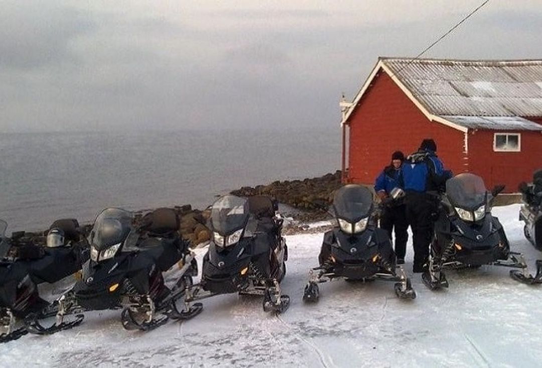 Six snowmobiles parked on top of a hill. Two men chatting among them. 