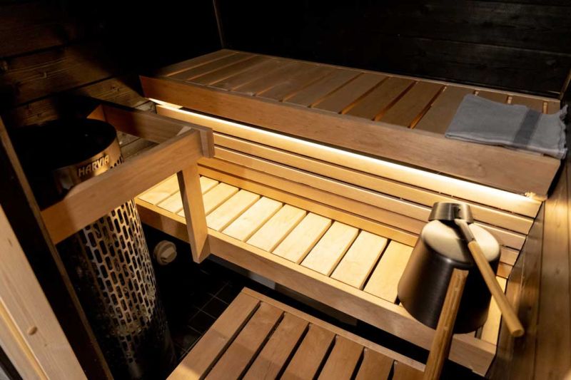 Sauna room with light coming under the seating made out of wood. Metallic, electrical  sauna stove in corner.