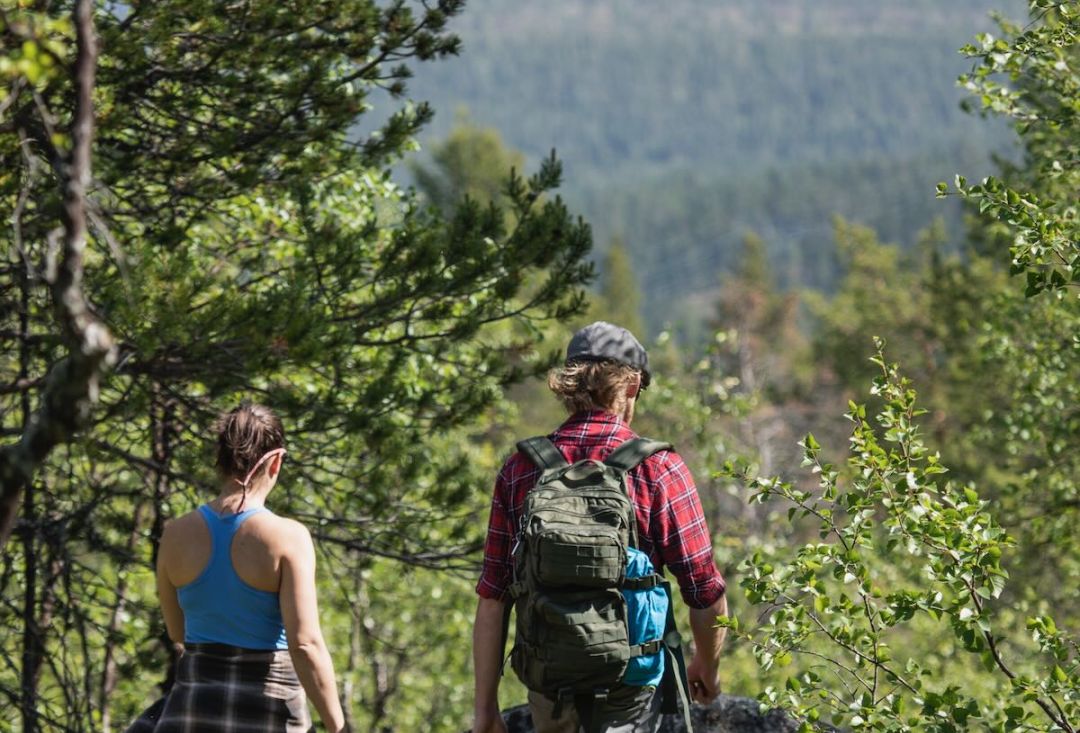 Pariskunta kävelee alas rinnettä kirkkaana kesäpäivänä, toisella reppu.
