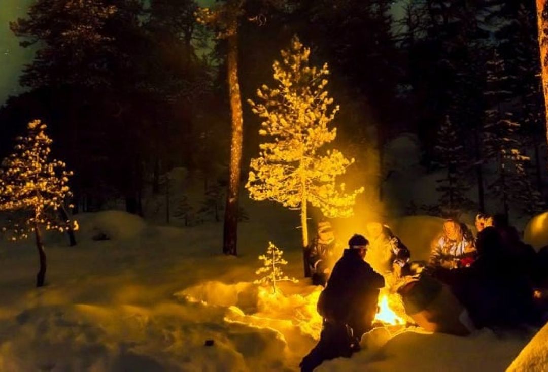 People sitting around bonfire in a dark forest filled with snowy trees.