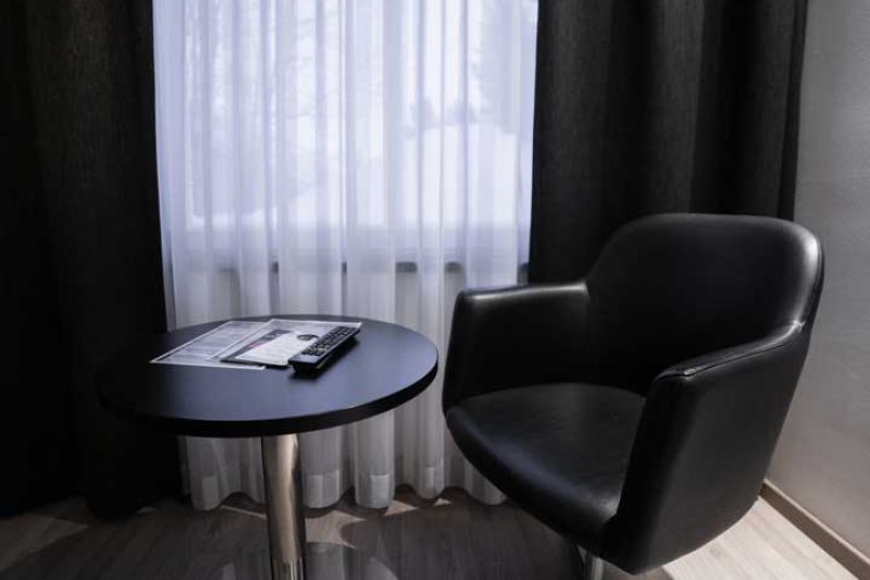 Little, black round table with leather armchair in front of window covered with light curtains.