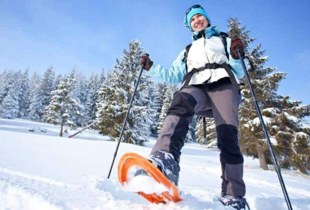 Hymyilevä nainen ottaa askelta lumikengällä. Taustalla havupuumetsä.
