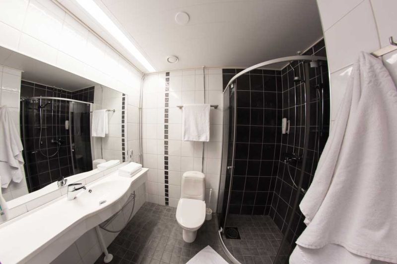 Vaalea kylpyhuone yhdellä mustalla seinällä. Wc-istuin, pitkä allaspöytä ja kulmasuihkukaappi.