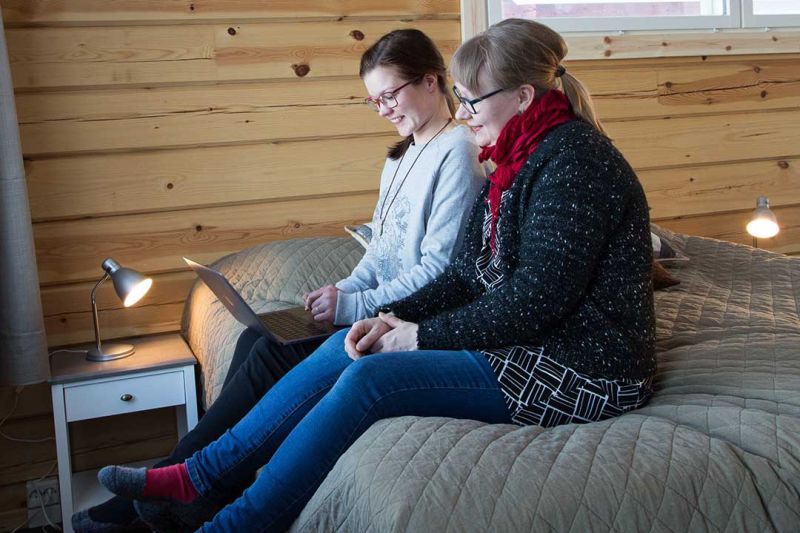 Kaksi naista istuu sängyllä katsellen kannettavaa tietokonetta. Sängyn vierellä pieni yöpöytä valaisimella.