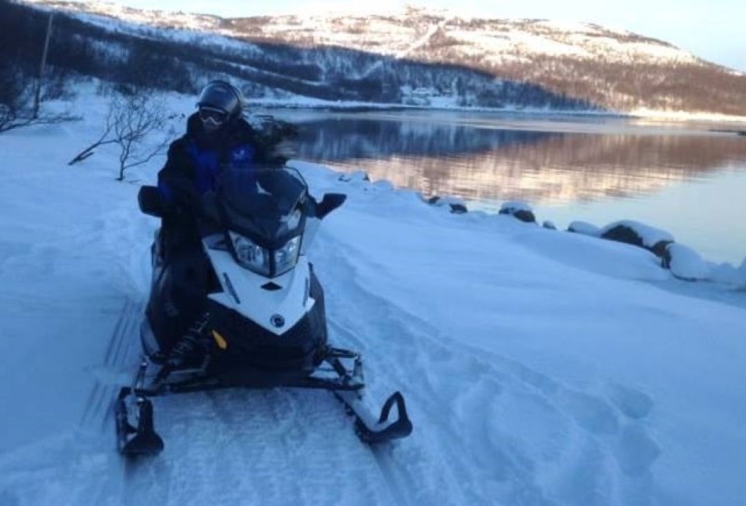A person sitting on a snowmobile by an unfrozen lake.
