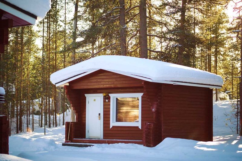 Mökki talvimaisemassa. Mökissä ulko-ovi, jonka vieressä suurehko ikkuna.