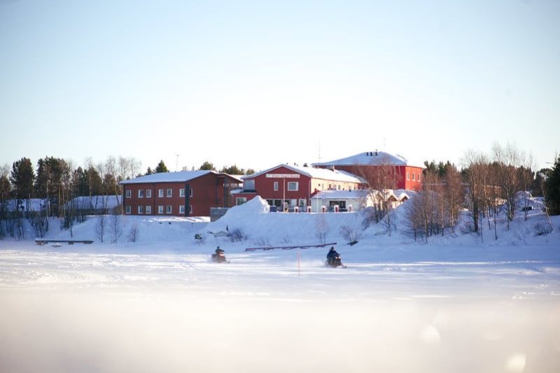 Hotelli järveltä päin katsottuna talvimaisemassa. Jäällä kaksi moottorikelkkailijaa.
